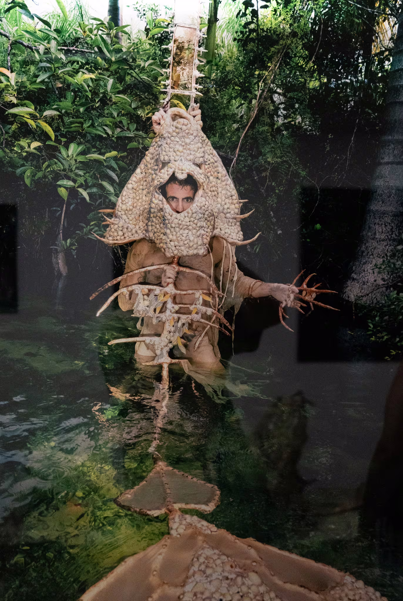 Person in elaborate shell and twig costume in lush green setting, partially submerged in water, creating a surreal and organic visual effect.