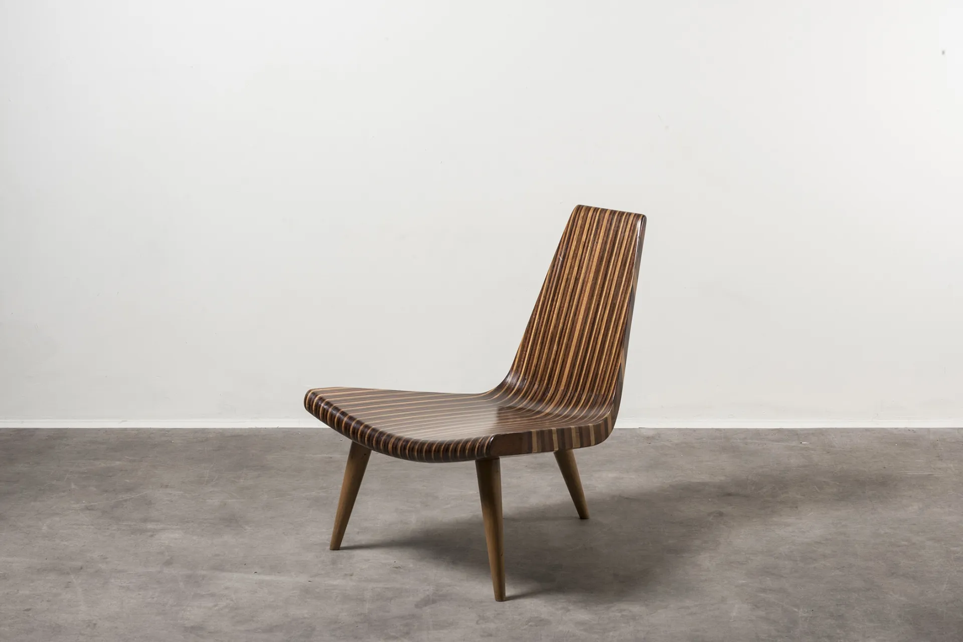 Low lounge chair with a striped wood-veneer seat and curved backrest on tapered wooden legs, set against a white wall and grey concrete floor.