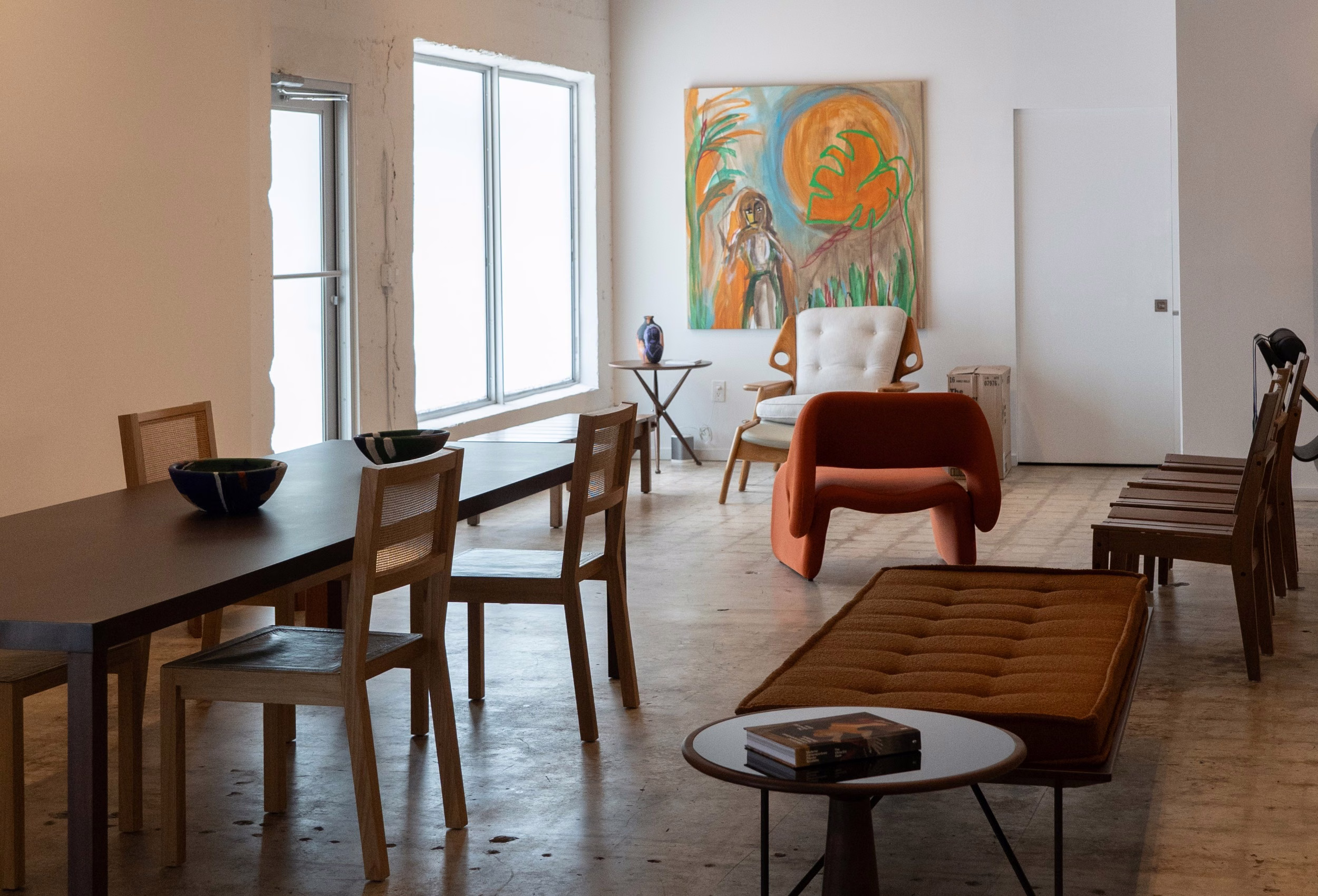 A dark wood dining table with cane-back chairs sits near a tufted daybed, an orange armchair and a bold painting in a bright, white-walled loft.