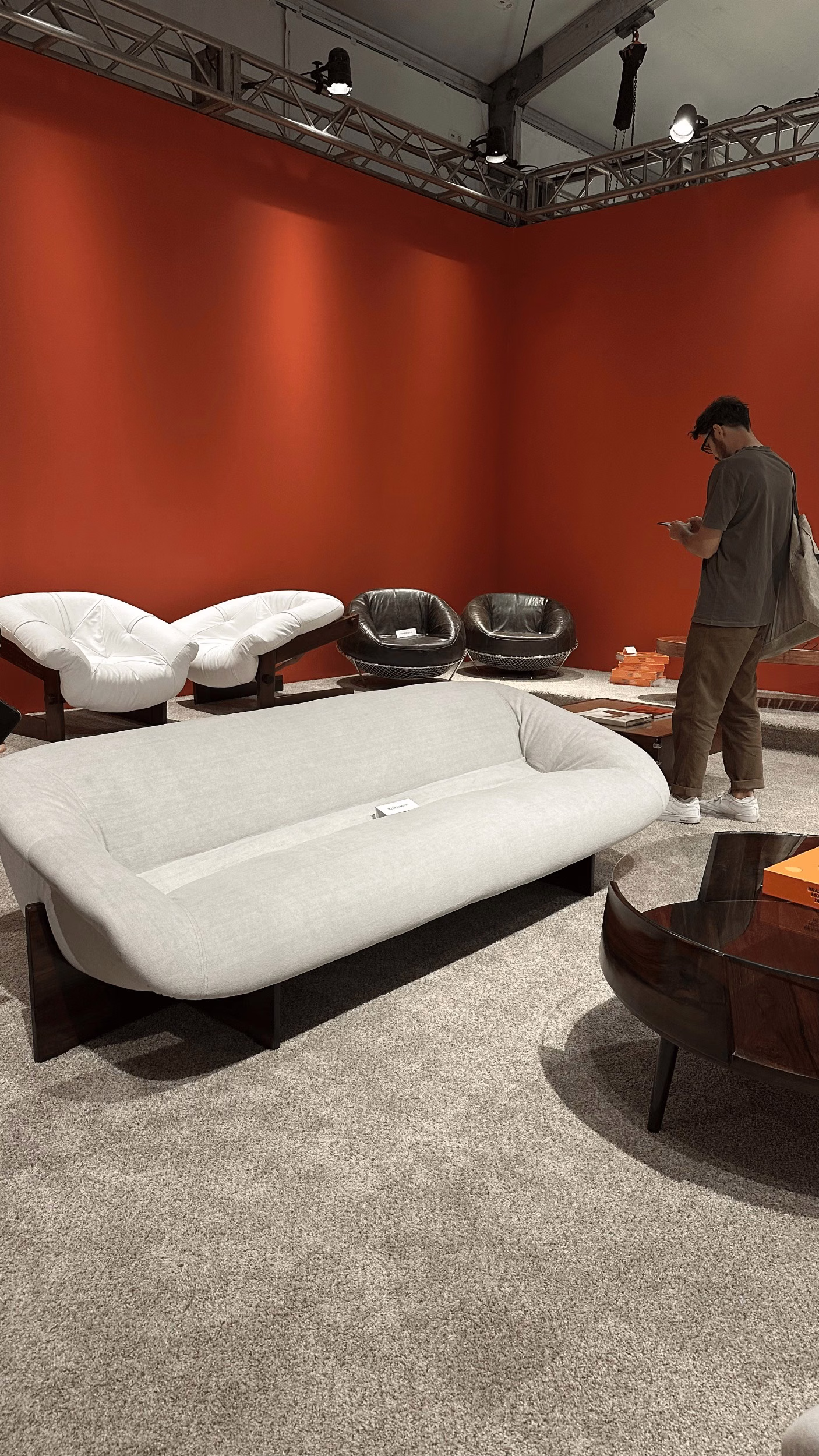 Rounded fabric sofa and leather armchairs on grey carpet in a showroom with burnt-orange walls and exposed ceiling trusses.