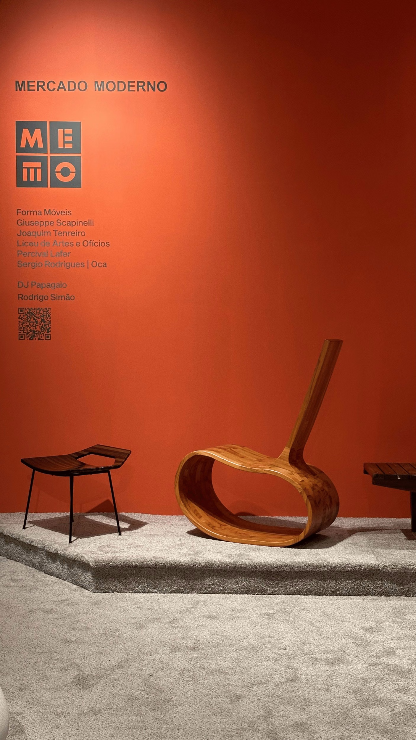 Sculptural bent-wood chair with a looping curved frame stands beside a slim dark wooden stool on a grey platform against a burnt-orange gallery wall.