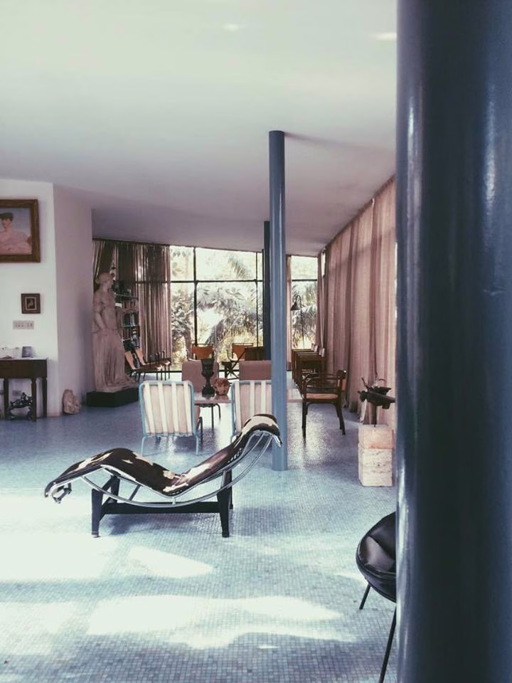 A cowhide chaise longue and leather chair sit on a mosaic-tiled floor beside slender columns, framed chairs, a stone sculpture and sheer curtains filtering daylight.
