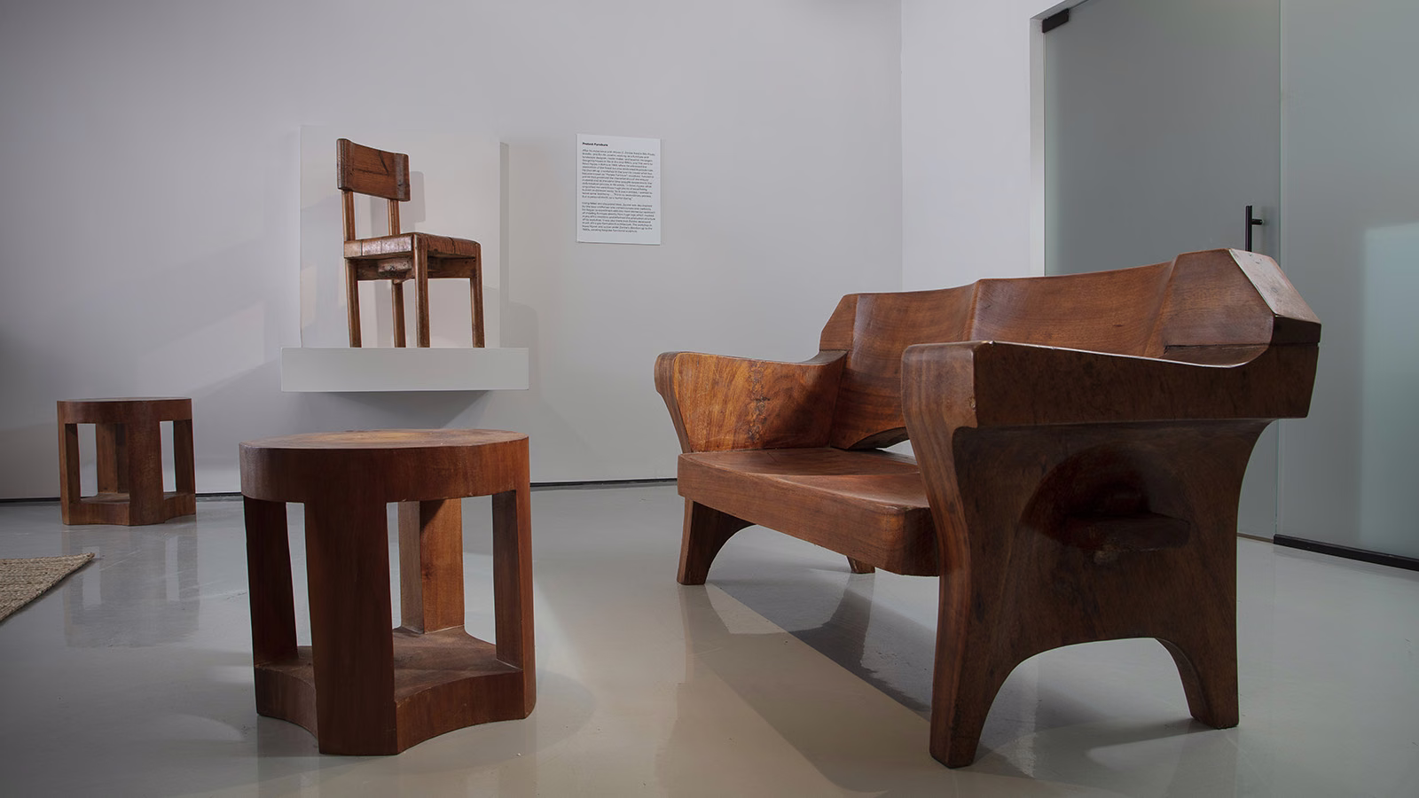 A sculptural wooden bench with curved legs sits beside two carved stools in a white gallery room, a small chair mounted on a wall shelf behind. José Zanine Caldas, R and company gallery