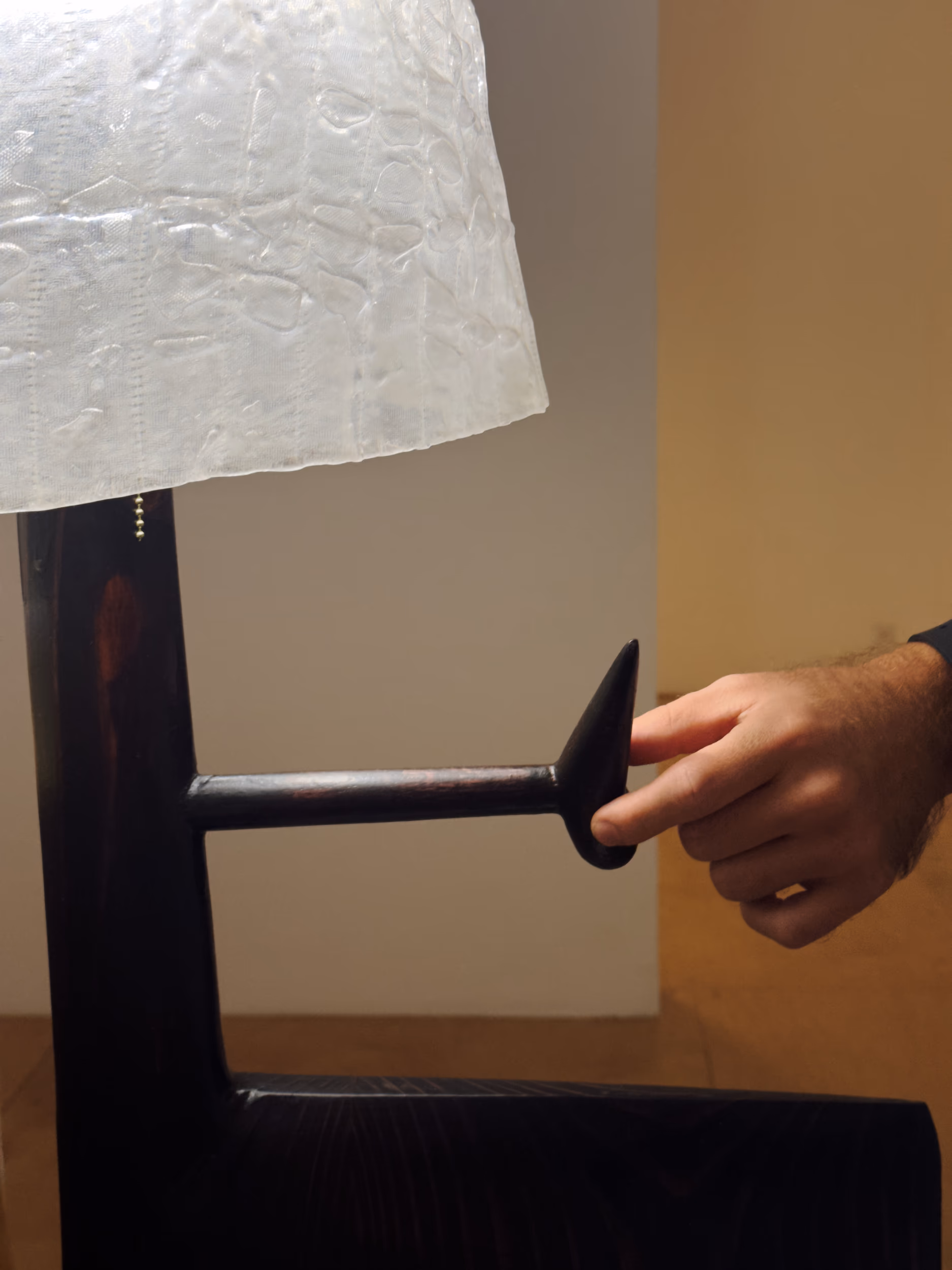A dark wooden lamp with paper shade and turned-wood arm being touched by a hand on a side table.