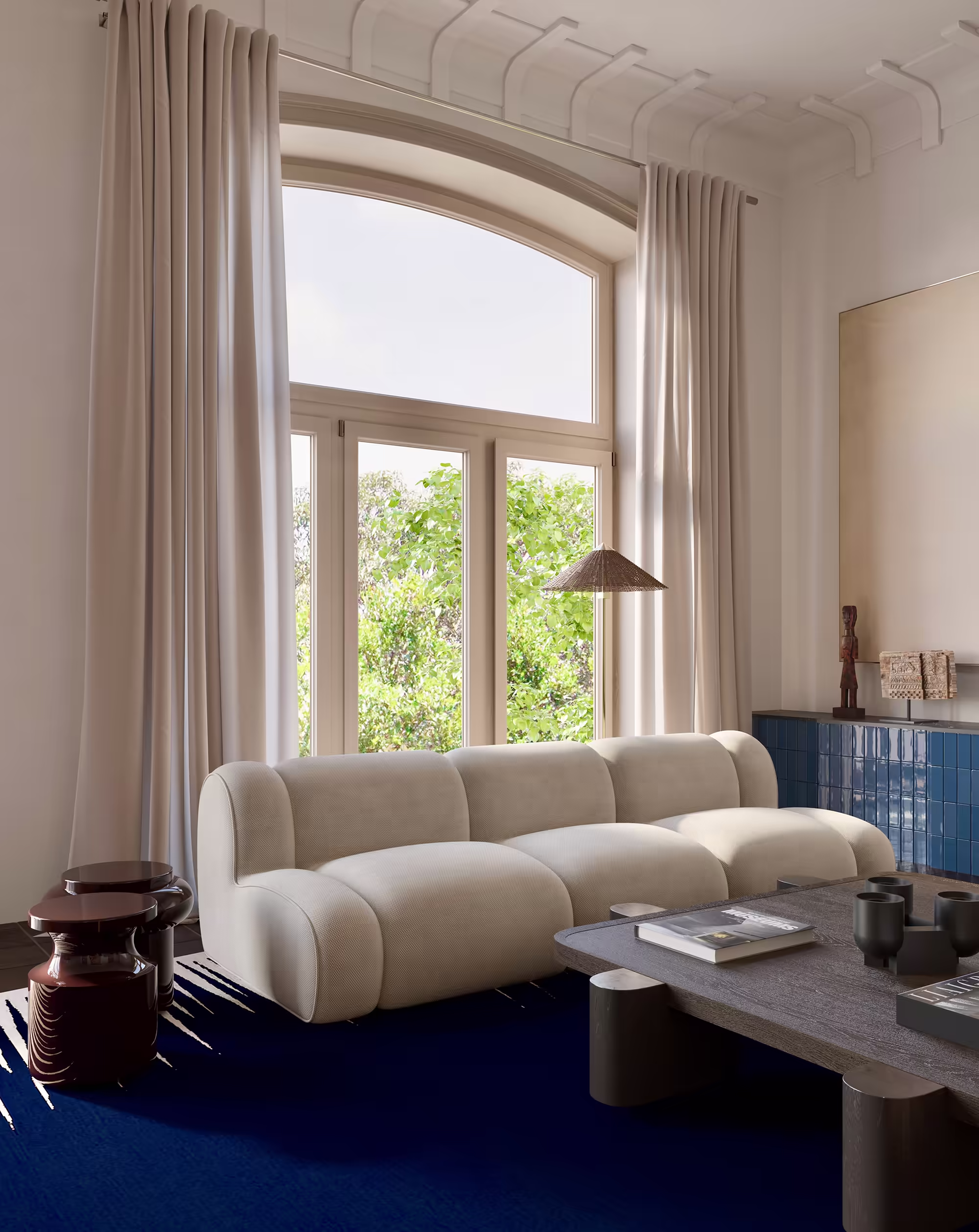 Bright living room with a sculptural white sofa Popus Edition Invisible collection and a dark wood and ceramic custom coffee table. Large arched window with creamy drapes frames a view of lush greenery. Deep blue rug by Atelier Fevrier and glossy burgundy side tables by Delcourt Collection anchor the space with contrast and richness. Vintage Paavo Tynnel floor lamp from 1stdibs.