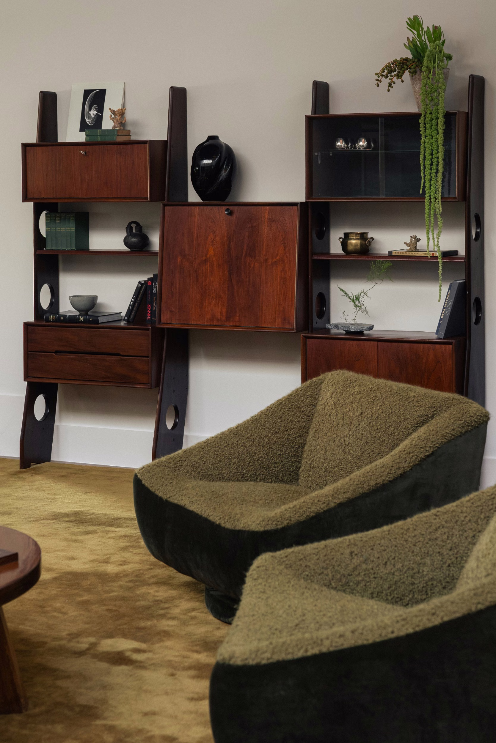Mid-century rosewood wall unit with cabinets, shelves and books beside two olive-green bouclé chairs on a mustard rug in a warm-lit room.