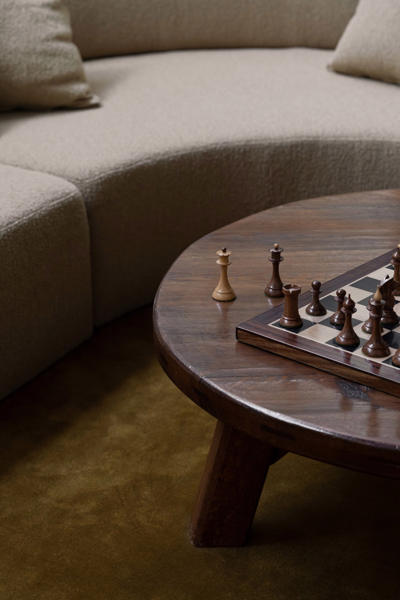Round dark-wood coffee table with a chess set mid-game beside a curved cream bouclé sofa on an olive-green rug in soft light.