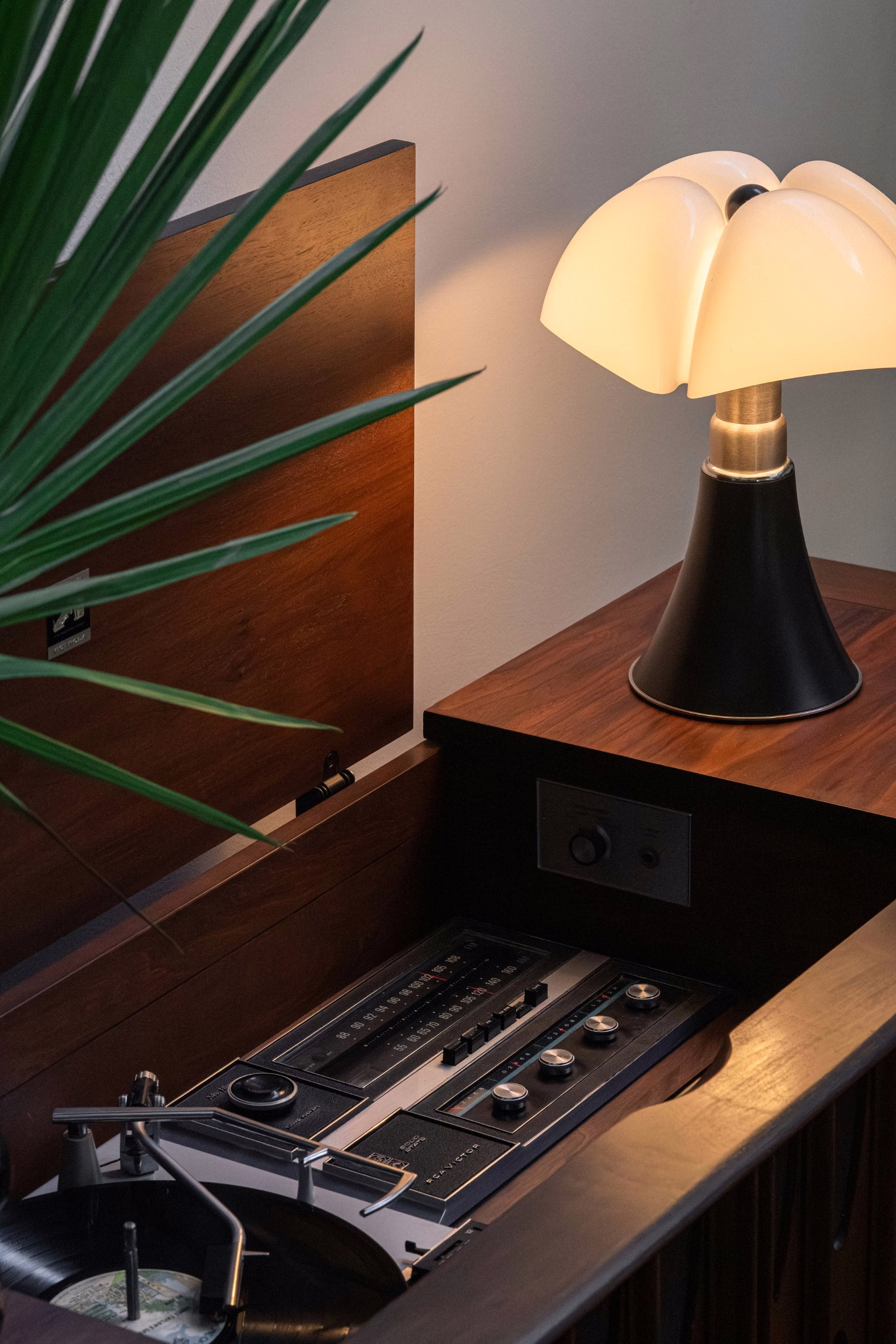 Warm-lit mushroom lamp with white shade and black base atop a walnut cabinet housing a vintage turntable, with green palm fronds in the foreground.