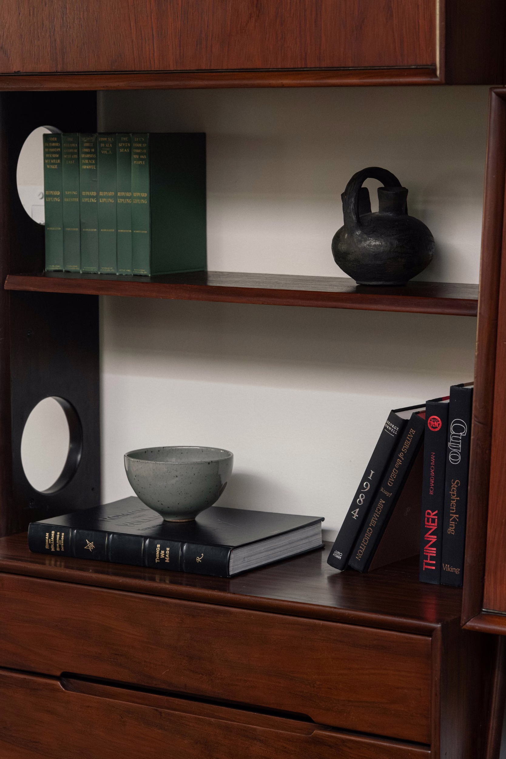 Dark wood bookcase with green cloth-bound books, a black ceramic vessel, grey speckled bowl, and stacked black books against a white wall.
