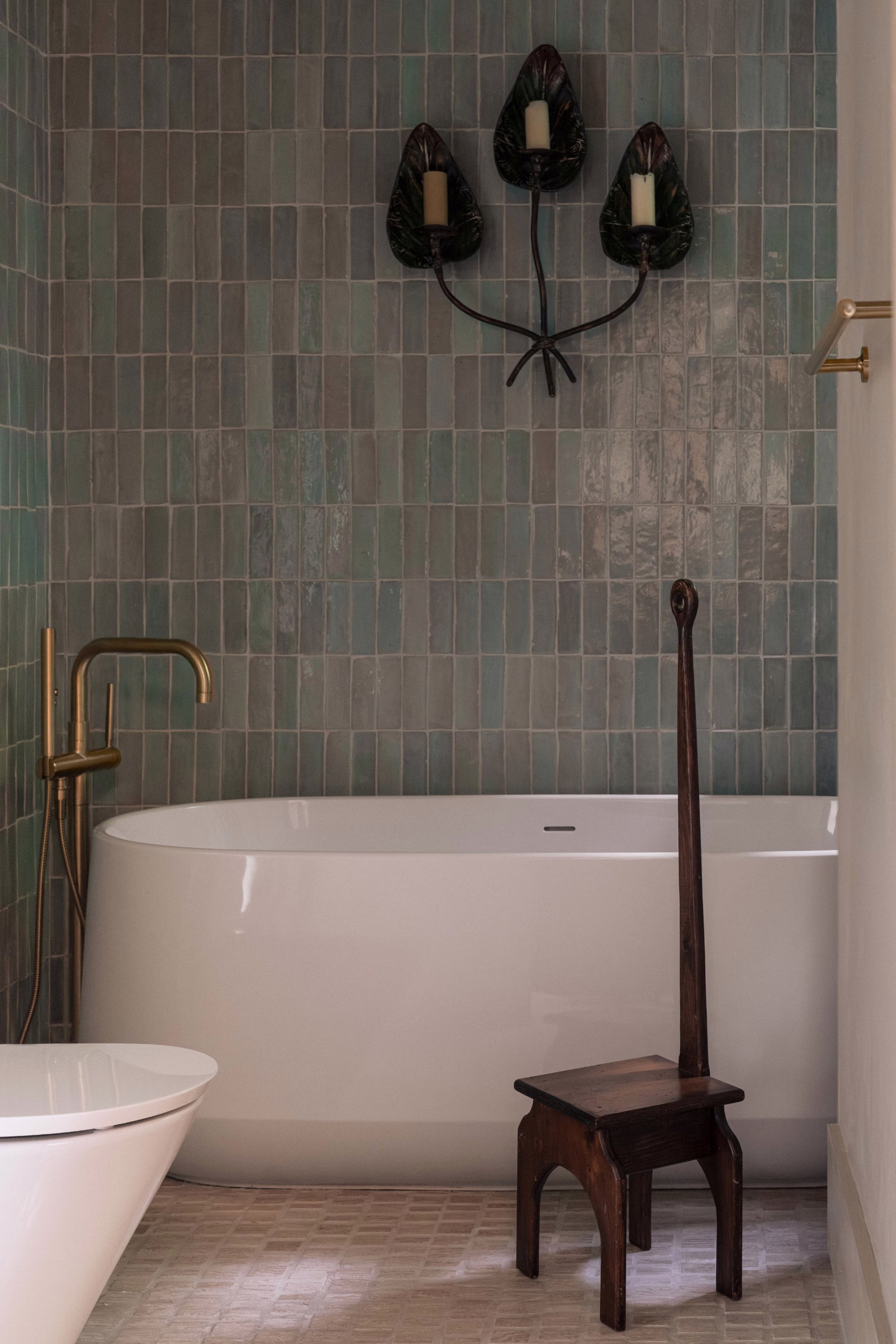 Freestanding white bathtub with brass floor-mounted tap against green vertical tiles, a dark wooden stool and leaf-shaped candle sconce above.