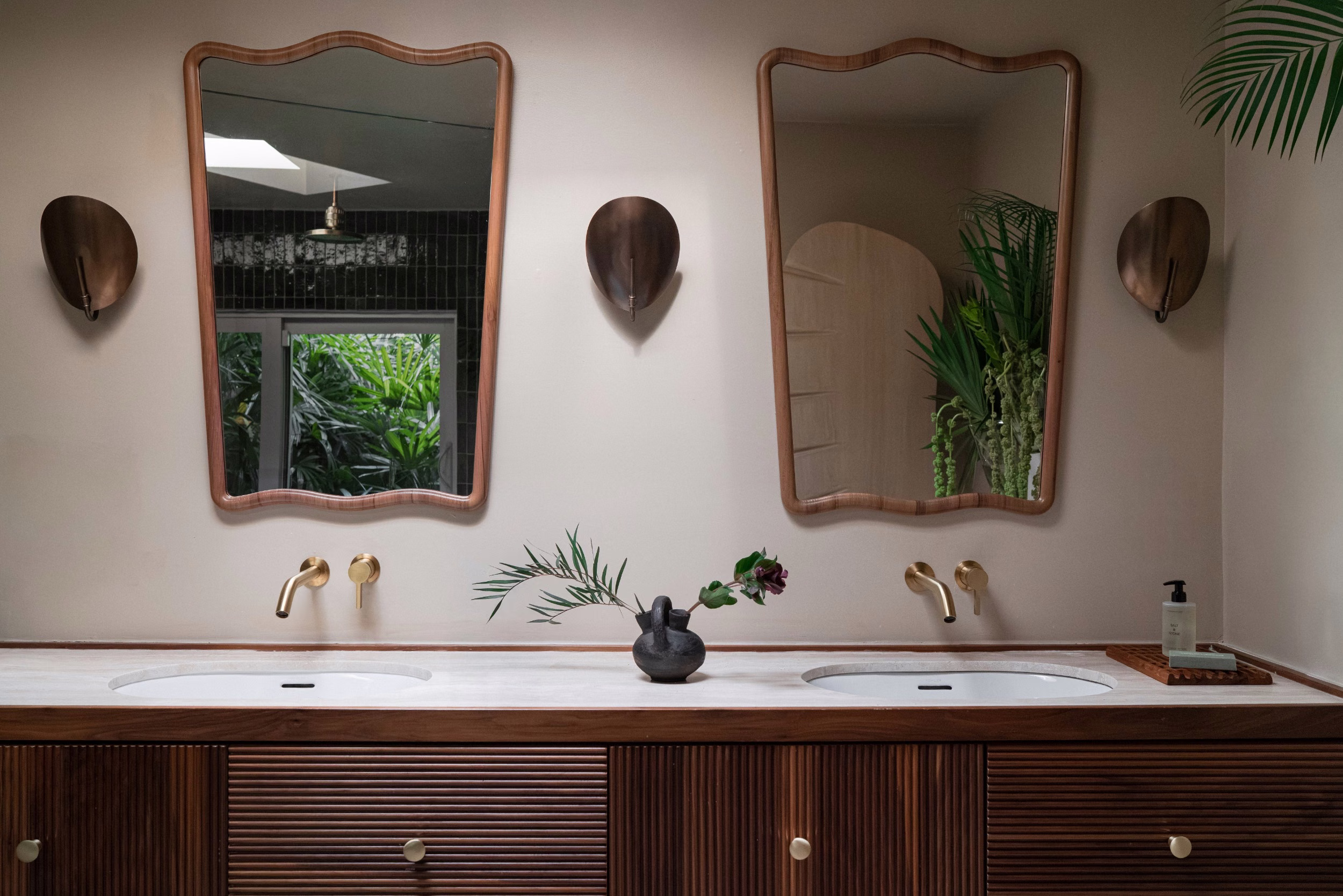 Double vanity with fluted walnut cabinetry, stone top, brass wall-mounted taps and sconces, twin wavy-framed mirrors, and a vase of foliage against beige walls.