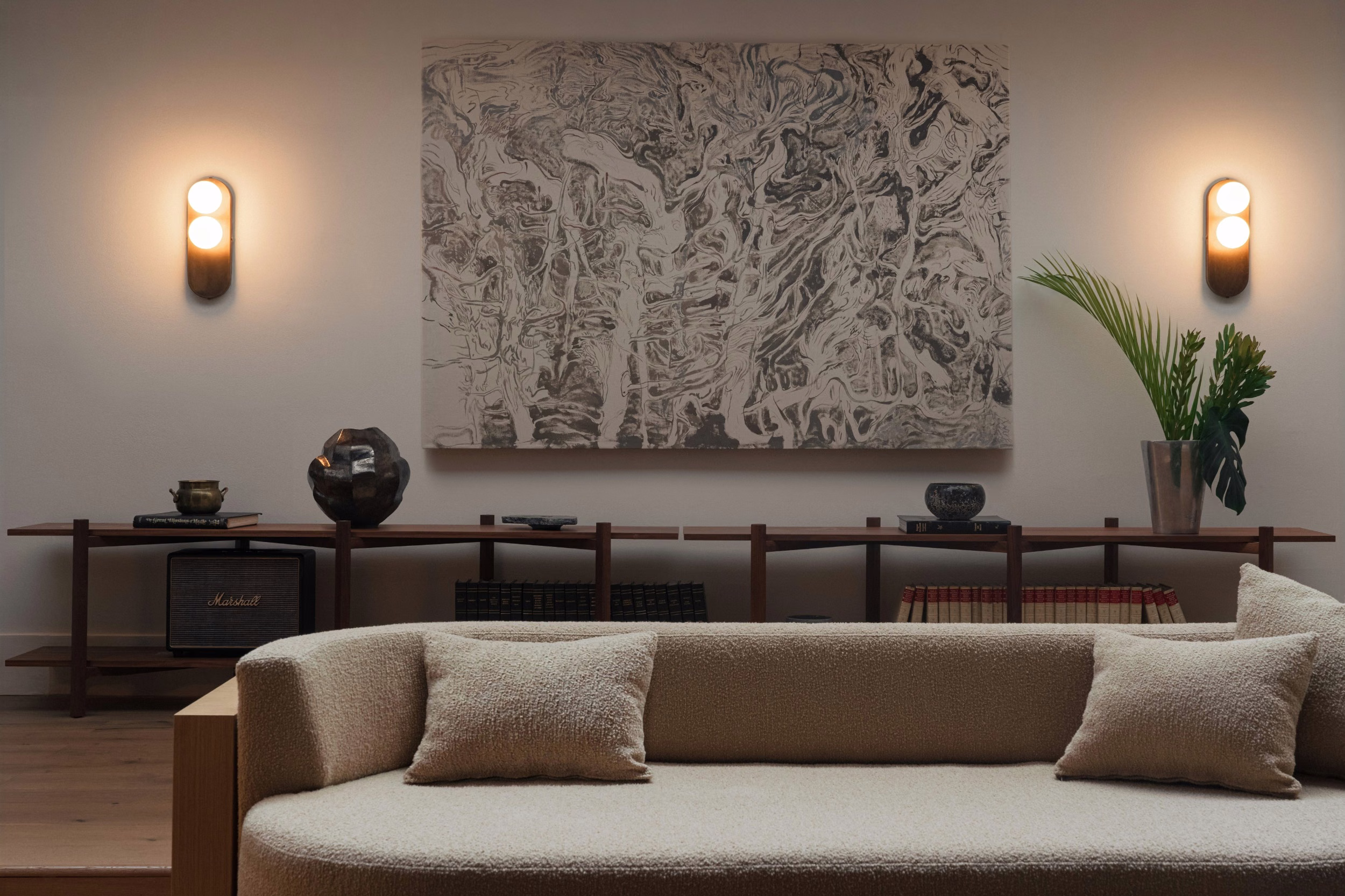 Cream bouclé sofa with cushions before a low wooden console and abstract black-and-white artwork, flanked by two oval wall sconces with warm light.