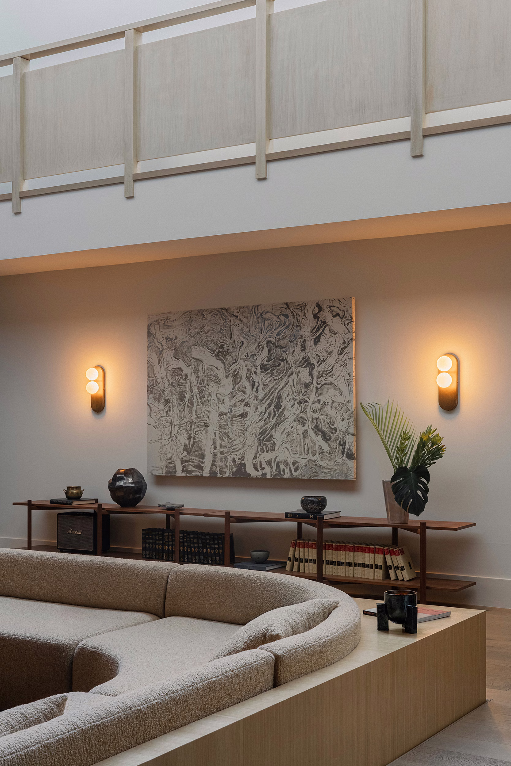 Curved cream bouclé sofa in a double-height room with an Kristina Aksentova’s artwork, two oval wall sconces, and a low walnut console holding books and plants.