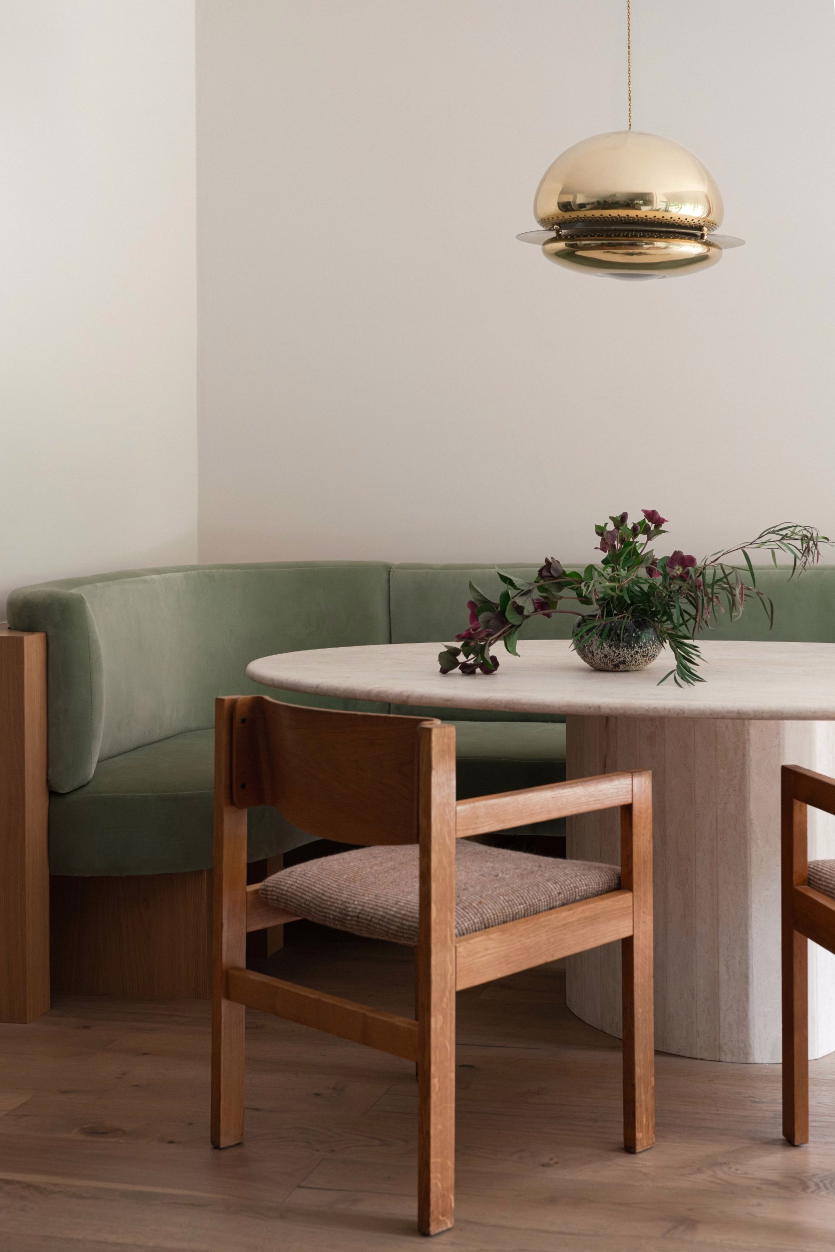Round travertine table with oak chairs beside a sage green velvet banquette, a brass pendant light above and a dark floral arrangement in a glass vase.