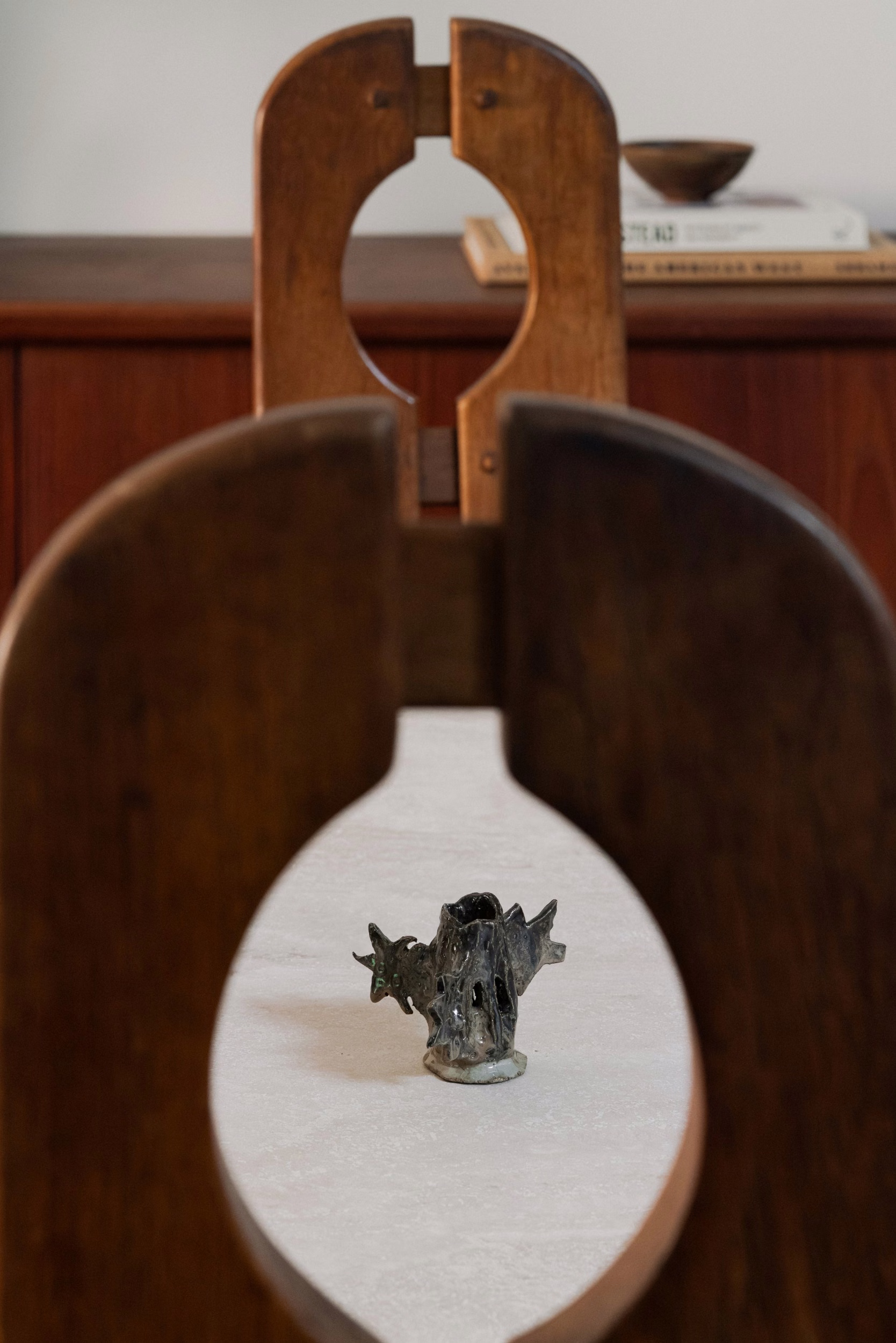 Small dark ceramic sculpture with bronze glaze on a pale stone surface, framed through the cutout backs of two wooden chairs, with a wood cabinet behind.